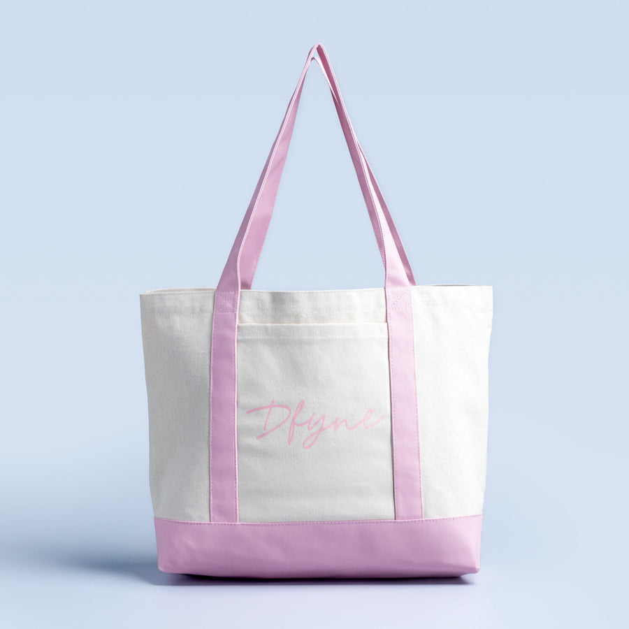 White canvas tote bag with lavender handles and bottom trim, cursive logo on front